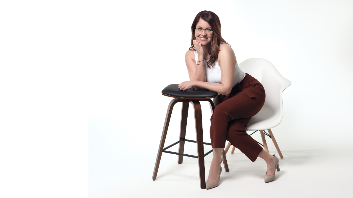 A woman in a white top and burgundy pants sitting on a white chair, leaning on a stool