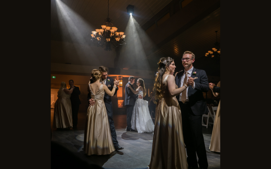 Couples dancing at a wedding with bride and groom in the center, illuminated by narrow spotlights.